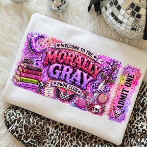 May include: A white sweatshirt featuring a vibrant graphic for the "Morally Gray Book Club." The design includes text, a ticket, a sword, a snake, and a stack of books. The color palette is primarily purple, pink, and white.