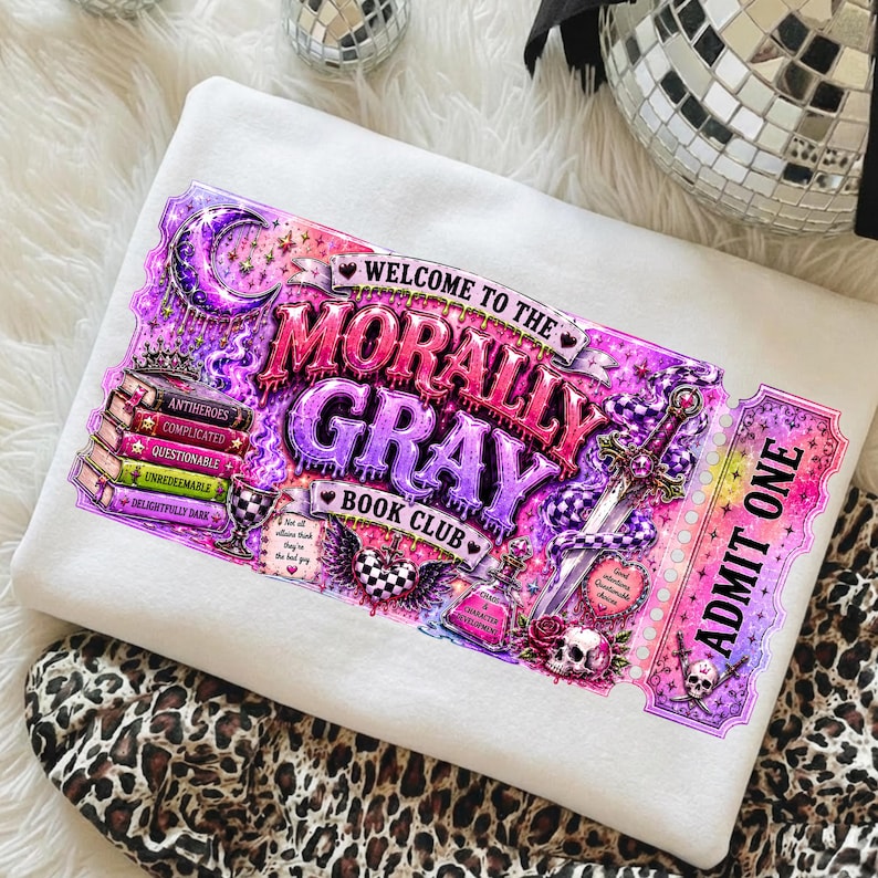 May include: A white sweatshirt featuring a vibrant graphic for the "Morally Gray Book Club." The design includes text, a ticket, a sword, a snake, and a stack of books. The color palette is primarily purple, pink, and white.
