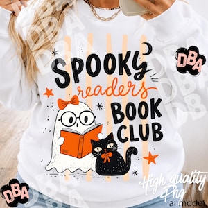 May include: A white long-sleeve sweatshirt featuring a graphic print with the text "SPOOKY readers BOOK CLUB". The design includes a white ghost wearing glasses and an orange bow, reading an orange book, next to a black cat with an orange bow.