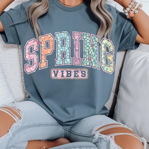 May include: A blue-grey t-shirt with the words "SPRING VIBES" in a floral design. The letters are filled with a variety of flowers in pink, orange, blue, and yellow. The shirt is a casual style.