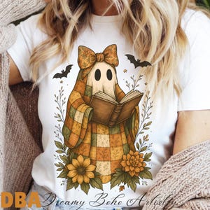 May include: White t-shirt featuring a whimsical ghost character reading a book. The ghost is draped in a plaid blanket and adorned with a bow. The design includes sunflowers, bats, and the text "Dreamy Boho Artistry".