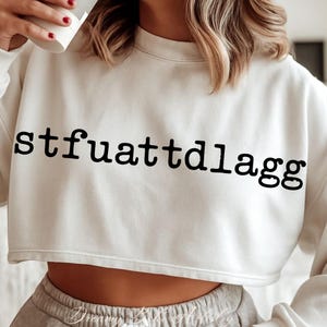 Puede incluir: Sudadera corta de color crema con el texto negro "stfuattdIagg" en negrita en la parte delantera. La sudadera tiene cuello redondo y mangas largas. El dobladillo inferior est&aacute; cortado justo por encima de la cintura.