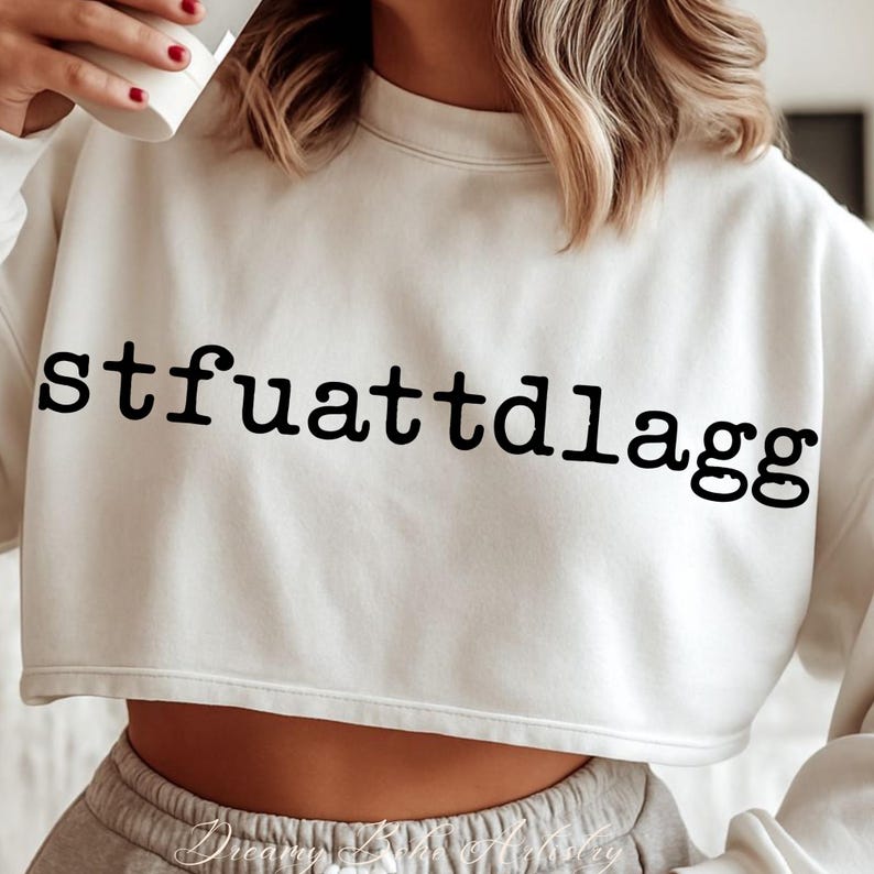 Puede incluir: Sudadera corta de color crema con el texto negro "stfuattdIagg" en negrita en la parte delantera. La sudadera tiene cuello redondo y mangas largas. El dobladillo inferior est&aacute; cortado justo por encima de la cintura.