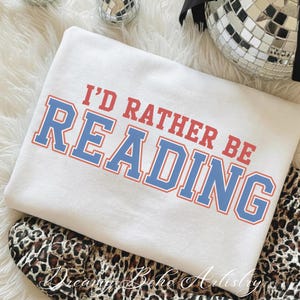 May include: White sweatshirt with the text "I'D RATHER BE READING" in red and blue lettering. The sweatshirt is folded and resting on a leopard print fabric. Decorative disco balls are in the background.