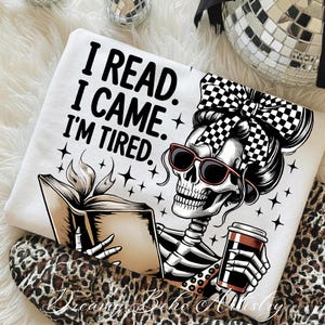 May include: White sweatshirt with the text "I READ. I CAME. I'M TIRED." featuring a skeleton wearing sunglasses and a checkered bow, reading a book and holding a coffee cup. The design includes star accents.