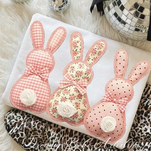 May include: A white cushion decorated with three appliqued bunny rabbits. Each bunny has a different pattern: pink gingham, floral, and pink polka dots. They have pink bows and white rosebud tails. The cushion is on a leopard print fabric.