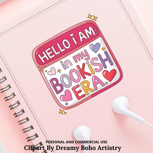 Op de afbeelding: Een roze notitieboek met een spiraalbinding en een sticker met de tekst "HELLO I AM in my BOOKISH ERA" in verschillende kleuren. De sticker heeft harten en sterren. Witte oortelefoons liggen op het roze oppervlak.