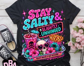 Puede incluir: Una camiseta negra con un estampado gráfico que incluye el texto STAY SALTY & SLIGHTLY UNHINGED y BEACH SNACKS & BAD DECISIONS. El diseño muestra una calavera con gafas de sol en forma de corazón, una bebida con calavera, patatas y flores de hibisco.