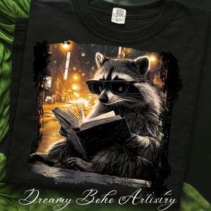 May include: Black t-shirt featuring a raccoon wearing sunglasses and reading a book. The raccoon is set against a city street scene with warm, golden lighting. The words "Dreamy Boho Artistry" are at the bottom.