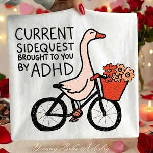 May include: White t-shirt with a whimsical illustration of a goose riding a bicycle with a basket of flowers. The text reads "CURRENT SIDEQUEST BROUGHT TO YOU BY ADHD". The design is in black and pink.
