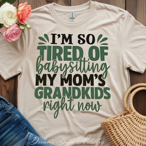 Può includere: T-shirt color crema con la scritta "I'M SO TIRED OF babysitting MY MOM'S GRANDKIDS right now" in lettere verdi e nere. La maglietta è esposta su una superficie di legno con fiori e un cestino di vimini.