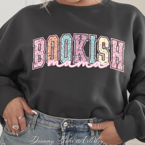 May include: Dark gray sweatshirt with the text "BOOKISH mama" in a floral design. The word "BOOKISH" is in a collegiate style font with a pink outline. The word "mama" is in a cursive font. The sweatshirt is a casual style.