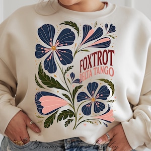 May include: A cream-colored sweatshirt with a floral design in shades of blue, green, and pink. The text "FOXTROT DELTA TANGO" is printed in red and burgundy.
