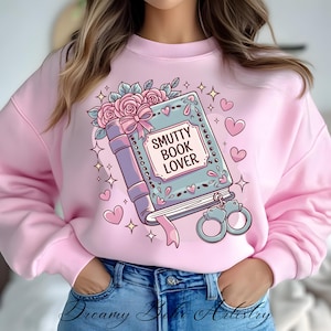 May include: Pink sweatshirt with a graphic of a blue book with the text "SMUTTY BOOK LOVER" and a pair of handcuffs. The book is decorated with pink flowers and a pink bow.