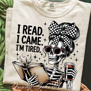 May include: Cream-colored t-shirt featuring a skeleton with sunglasses and a checkered bow, reading a book and holding a coffee cup. The shirt has the text "I READ. I CAME. I'M TIRED." in black. The design includes star accents.