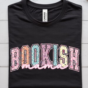 May include: Black t-shirt with the text "BOOKISH mama" in a colorful, floral design. The word "BOOKISH" is in a bold, block letter style, and "mama" is in a cursive font. The shirt is made of a soft material.