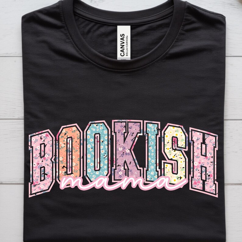 May include: Black t-shirt with the text "BOOKISH mama" in a colorful, floral design. The word "BOOKISH" is in a bold, block letter style, and "mama" is in a cursive font. The shirt is made of a soft material.