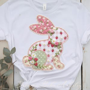 May include: White t-shirt featuring a decorative bunny applique. The bunny is made of various patterned fabrics, including pink floral, gingham, and floral prints. It is adorned with small flowers, strawberries, and a pearl-like trim. The text "Dreamy Behe Artistry" is at the bottom.