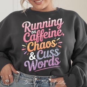 May include: A dark grey sweatshirt with the phrase "Running on Caffeine, Chaos & Cuss Words" in a colourful, playful font. The design features pink, orange, and purple hues, with a whimsical style. The sweatshirt is a casual, comfortable garment.