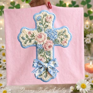 May include: Embroidered cross with a floral design in pink, blue, and cream colors. The cross is outlined in light blue and features a bow at the bottom. The background is a soft pink.