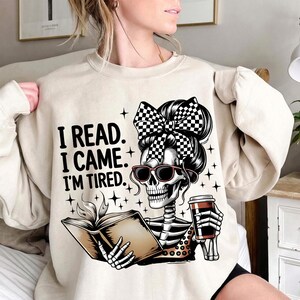 May include: Beige sweatshirt with the text "I READ. I CAME. I'M TIRED." features a skeleton graphic wearing sunglasses and a checkered bow, reading a book and holding a coffee cup. The design includes star accents.