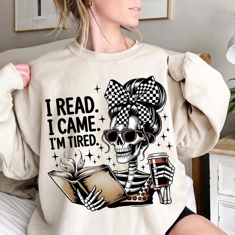 May include: Beige sweatshirt with the text "I READ. I CAME. I'M TIRED." features a skeleton graphic wearing sunglasses and a checkered bow, reading a book and holding a coffee cup. The design includes star accents.