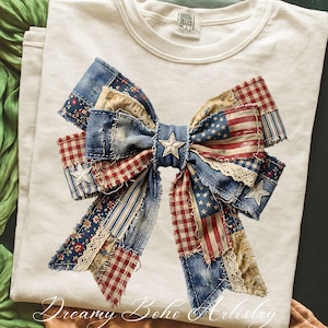 May include: Cream-coloured t-shirt with a large patchwork bow design. The bow incorporates denim, red and white gingham, floral, and American flag patterns. A silver star embellishes the centre. The text "Dreamy Boho Artistry" is at the bottom.