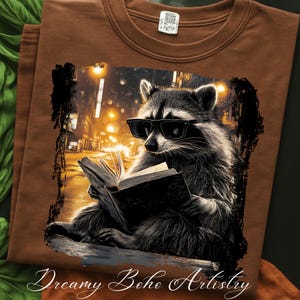 May include: Brown t-shirt featuring a raccoon wearing sunglasses and reading a book. The raccoon is set against a city street scene with warm, golden lighting. The text "Dreamy Boho Artistry" is at the bottom.