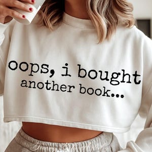 Puede incluir: Sudadera corta de color crema con el texto "oops, i bought another book..." en negro. La sudadera tiene cuello redondo y mangas largas. El texto está en una fuente de estilo máquina de escribir.