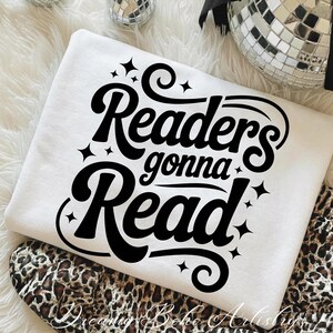 K&ouml;nnte beinhalten: Ein wei&szlig;es Sweatshirt mit dem schwarzen Schriftzug "Readers gonna Read" in einer stilisierten Schriftart mit Sternen. Das Sweatshirt liegt gefaltet auf einem Stoff mit Leopardenmuster.