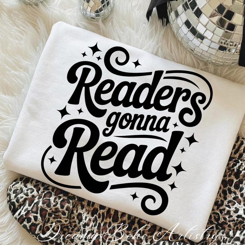K&ouml;nnte beinhalten: Ein wei&szlig;es Sweatshirt mit dem schwarzen Schriftzug "Readers gonna Read" in einer stilisierten Schriftart mit Sternen. Das Sweatshirt liegt gefaltet auf einem Stoff mit Leopardenmuster.