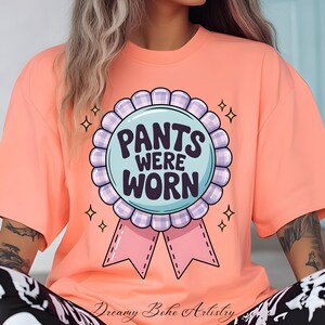Puede incluir: Camiseta color melocotón con un gráfico redondo que dice "PANTS WERE WORN". El gráfico es un círculo azul claro con un borde a cuadros morado y blanco, y una cinta rosa. La camiseta tiene mangas cortas.