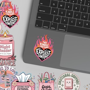 May include: A collection of colorful stickers with book-themed designs. One sticker features a pink candle with the text "Night Reader." Another sticker has the text "This Kindle Contains Explicit Content" in a heart shape. A laptop is in the background.