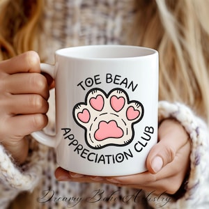Puede incluir: Taza de cerámica blanca con una ilustración negra y rosa de una huella de pata con el texto "Toe Bean Appreciation Club".