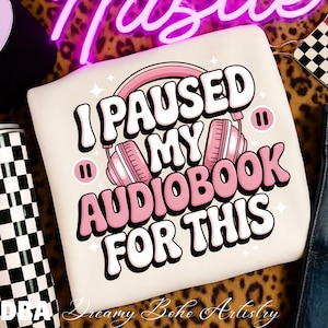 May include: Cream-colored sweatshirt with the text "I PAUSED MY AUDIOBOOK FOR THIS" in white and pink. The design includes pink headphones and pause symbols. The sweatshirt is likely made of a soft material.