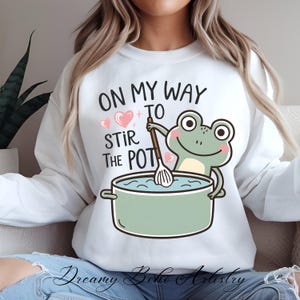Könnte beinhalten: Weißer Pullover mit einem grünen Frosch, der einen Schneebesen hält und einen Topf mit Wasser umrührt. Der Text auf dem Pullover lautet "On my way to stir the pot".