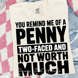 Könnte beinhalten: Cremefarbenes T-Shirt mit schwarzem Aufdruck: "YOU REMIND ME OF A PENNY TWO-FACED AND NOT WORTH MUCH." Das Shirt ist gefaltet und liegt auf einer rosa-weiß karierten Oberfläche und einer Jeans.