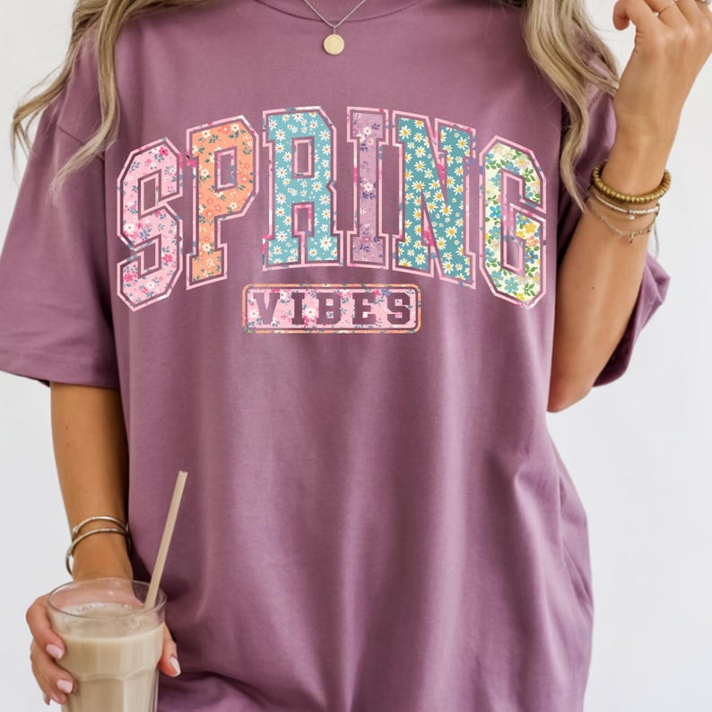 May include: A mauve-colored t-shirt with the words "SPRING VIBES" in a floral design. The word "SPRING" is in a collegiate-style font, with each letter featuring a different floral pattern. The word "VIBES" is in a rectangular box.
