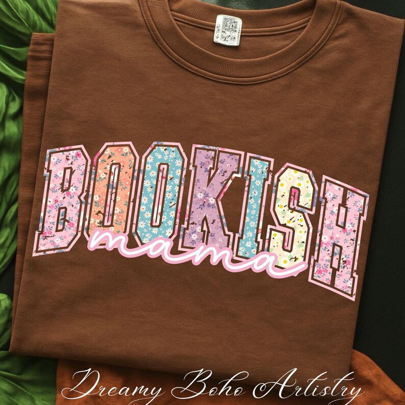 May include: A brown t-shirt with the words "BOOKISH mama" in a floral design. The text is in a collegiate style font with a pink outline. The shirt is folded and displayed on a dark surface.