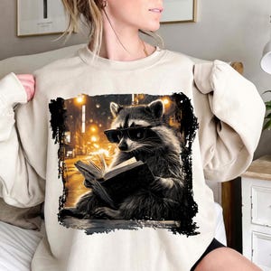 May include: Beige sweatshirt featuring a graphic of a raccoon wearing sunglasses and reading a book. The raccoon is set against a blurred city background with streetlights. The graphic is framed with a black border.