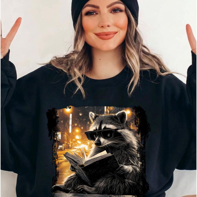 May include: Black sweatshirt featuring a raccoon wearing sunglasses and reading a book. The raccoon is set against a backdrop of a city street at night, with warm yellow streetlights. The design is a graphic illustration.
