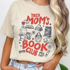 Op de afbeelding: Beige T-shirt met de rode tekst "Tired Mom's Book Club". Het ontwerp bevat illustraties van boeken, een leeslamp, een kat en andere boekgerelateerde items. De afbeelding is gedrukt op een shirt met korte mouwen.
