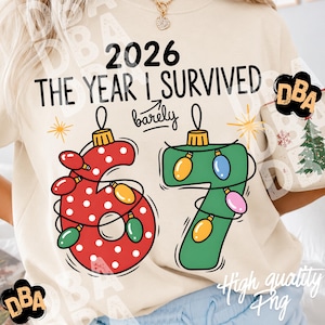 May include: A cream-colored t-shirt featuring the black text 2026 THE YEAR I SURVIVED barely, with a red polka-dot number 6 and a green number 7 shaped like Christmas ornaments, wrapped in colorful string lights and accented with yellow stars.