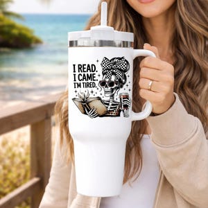 May include: White travel mug with a handle and a lid, featuring a skeleton graphic with sunglasses, a checkered bow, and the text "I READ. I CAME. I'M TIRED." The skeleton is reading a book and holding a coffee cup.