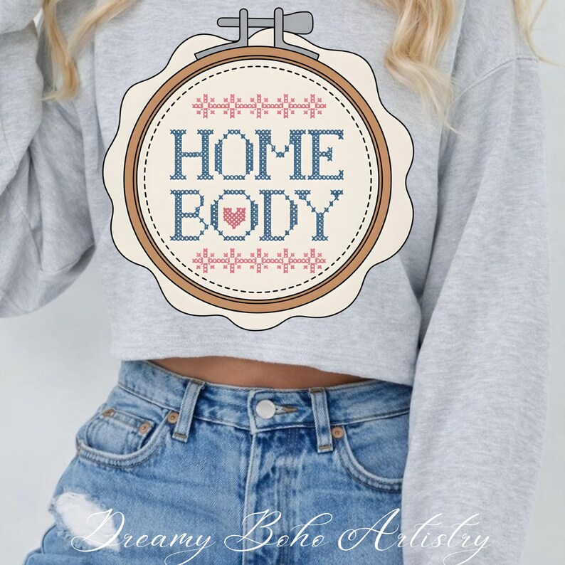 May include: A light grey sweatshirt with a cross-stitch design in an embroidery hoop. The design says "HOME BODY" in blue, with a pink heart. The sweatshirt is worn with blue denim jeans.