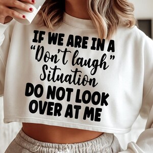 May include: Cream-colored cropped sweatshirt with the text "IF WE ARE IN A 'Don't laugh' Situation DO NOT LOOK OVER AT ME" in black. The sweatshirt is a casual style, suitable for everyday wear.