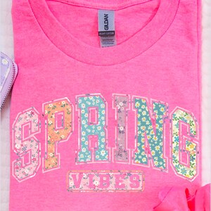 May include: A bright pink t-shirt with the word "SPRING" in a floral design across the front. Below the word "SPRING" is the word "VIBES" in a floral design. The shirt is made of heavy cotton.