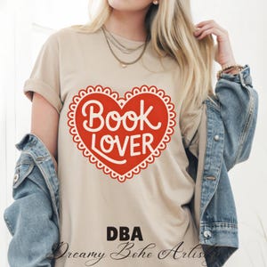 Puede incluir: Camiseta beige con un gran corazón rojo con borde festoneado. El diseño del corazón tiene las palabras "Book Lover" en escritura blanca. El texto "DBA Dreamy Boho Artis" está impreso debajo del corazón. La persona lleva una chaqueta vaquera.