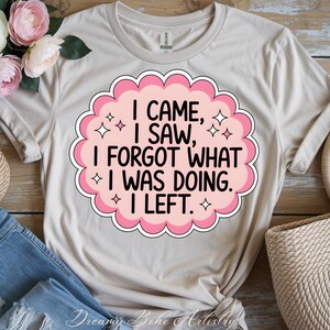 May include: A beige t-shirt with a pink and white graphic that reads "I CAME, I SAW, I FORGOT WHAT I WAS DOING. I LEFT." The design is surrounded by a pink cloud-like border with star accents. The shirt is folded on a wooden surface.