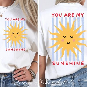 May include: White t-shirt with a yellow sun graphic and the text "YOU ARE MY SUNSHINE" in red. The sun has a smiling face and is set against blue vertical stripes. The design is on the front and back of the shirt.